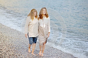 Redhead senior mother and her adult beautiful pregnant daughter are walking together on the sea shore