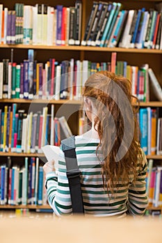 Redhead student reading book from shelf standing in library