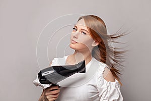 Redhead woman with blow drying hair holding hair dryer on gray wall studio background