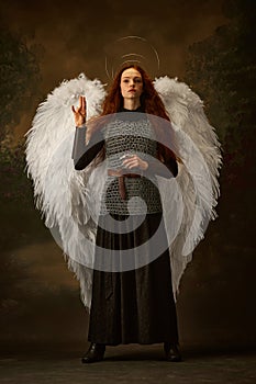 Redhead woman in chainmail and black skirt with large white wings and halo looks as angelic warrior against vintage