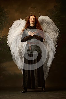 Redhead woman in chainmail and skirt with large white wings and halo looks as angelic warrior against vintage studio