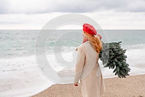 Redhead woman Christmas tree sea. Christmas portrait of a happy redhead woman walking along the beach and holding a