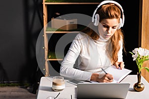 Redhead woman in headphones writing in notepad while watching webinar on laptop in cafe