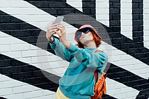 Redhead woman making duck face while taking a selfie photo outdoors. Emotional hipster fashion women in bright clothes