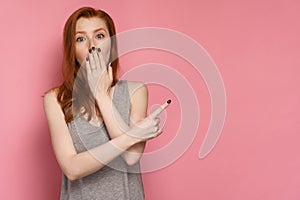 Redhead woman points her finger at blank space on the right of the photo covering her mouth with her hand in amazement