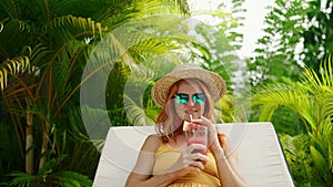 Redhead woman relaxes by poolside, sipping watermelon cocktail in sun, sunglasses. Leisure in tropical paradise, enjoys
