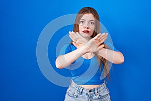 Redhead woman standing over blue background rejection expression crossing arms and palms doing negative sign, angry face