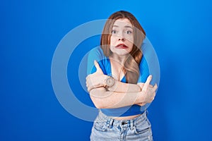 Redhead woman standing over blue background shaking and freezing for winter cold with sad and shock expression on face