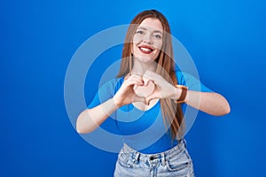 Redhead woman standing over blue background smiling in love doing heart symbol shape with hands