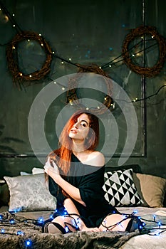 Redhead woman stay at cozy home alone to celebrate new year and lying on a gray bed with christmas lights and a garland.