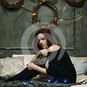Redhead woman stay at cozy home alone to celebrate new year and lying on a gray bed with christmas lights and a garland.