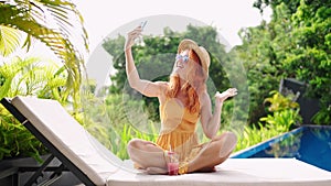 Redhead woman in summer dress lounges by poolside with tropical drink, captures sunny selfies using her smartphone