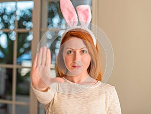 Redhead woman wearing easter rabbit ears at home with open hand doing stop sign with serious and confident expression, defense