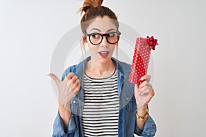 Redhead woman wearing glasses holding valentine gift over isolated white background pointing and showing with thumb up to the side