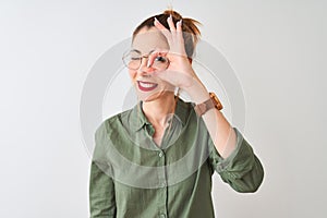 Redhead woman wearing green shirt and glasses standing over isolated white background doing ok gesture with hand smiling, eye
