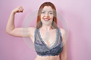 Redhead woman wearing lingerie over pink background strong person showing arm muscle, confident and proud of power