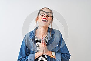 Redhead woman wearing striped t-shirt denim shirt and glasses over isolated white background begging and praying with hands