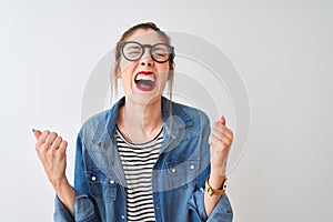 Redhead woman wearing striped t-shirt denim shirt and glasses over isolated white background very happy and excited doing winner