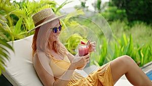 Redhead woman in yellow dress relaxes by tropical poolside, sips watermelon juice from glass. Sunhat, sunglasses, lush