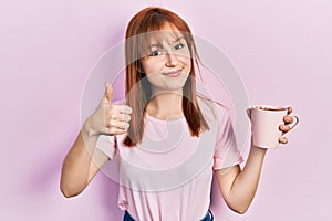 Redhead young woman drinking a cup coffee smiling happy and positive, thumb up doing excellent and approval sign