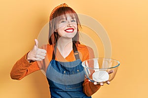 Redhead young woman holding bread dough smiling happy and positive, thumb up doing excellent and approval sign