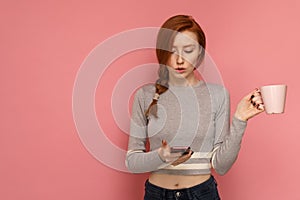 Redhead young woman holds a cup in one hand staring at her smartphone in other hand surprisingly