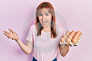 Redhead young woman showing fresh eggs celebrating achievement with happy smile and winner expression with raised hand