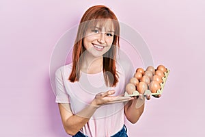 Redhead young woman showing fresh eggs looking positive and happy standing and smiling with a confident smile showing teeth
