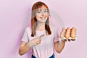 Redhead young woman showing fresh eggs smiling happy pointing with hand and finger