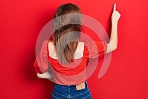 Redhead young woman wearing casual red t shirt posing backwards pointing ahead with finger hand