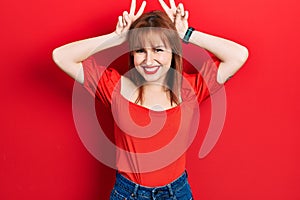 Redhead young woman wearing casual red t shirt posing funny and crazy with fingers on head as bunny ears, smiling cheerful