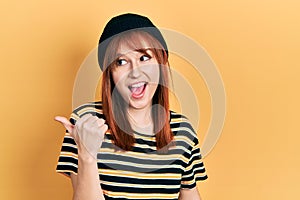 Redhead young woman wearing wool cap smiling with happy face looking and pointing to the side with thumb up