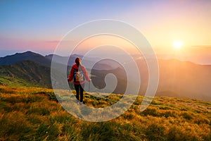 Redheaded girl athlete with a backpack and sticks stands on the green hillocks and looks at high mountain landscapes.