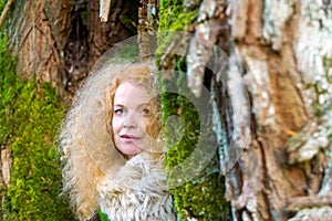 Redheaded woman in her fifties looks out between two moss-covered tree trunks, copy space