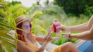 Redheaded woman relaxes by blue pool, receives vibrant watermelon cocktail. Luxury tropical leisure scene, breezy, sunny