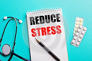 REDUCE STRESS written in a white notepad near a stethoscope, pens and pills on a light blue background. Medical concept