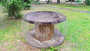 A reel made of wooden planks is located on a grassy lawn near trees, buildings and a walkway. Such reels are usually used to store