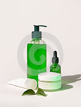 Refillable Cosmetic bottles, jar and tube near green leaves on white, bottles filled with green liquid