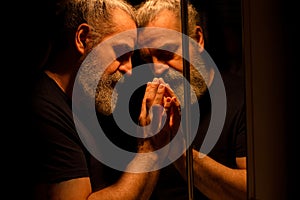 Reflection of bearded man in a dark, holding his head with his hands with painful expression