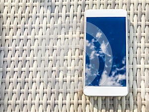 Reflection of blue sky with clouds in the phone-smartphone, which lies on a bright wicker table against the background. Blur and