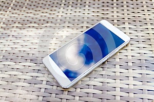 Reflection of blue sky with clouds in the phone-smartphone, which lies on a bright wicker table against the background. Blur and