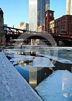 Reflection of buildings in water while bridge spans a frozen Chicago River with ice chunks.