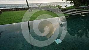 Reflection of fluffy clouds and palm trees in outside swimming pool in retro island villa yard. Evergreen grass and blue