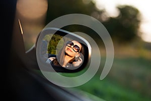 Reflection of a gorgeous young dark-haired girl in sunglasses in a side mirror of a car.