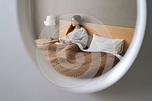 Reflection in mirror of cheerful young woman enjoy shopping online with credit card and mobile phone lying in bed at