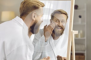 Reflection in mirror of funny adult bearded man who smears his face with moisturizer.