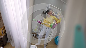 Reflection in mirror of joyful young woman dancing with broom imitating guitar playing. Wide shot portrait of cheerful