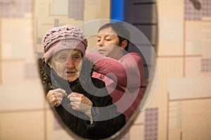 Reflection in the mirror, a woman helps put on her old mother`s coat