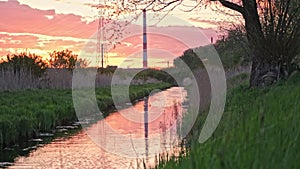 Reflection of Morning Sky with Distant Power Plant Refinery Tall Smokestack Chimney Cooling Tower in Narrow Creek Water