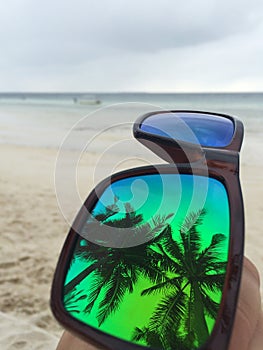Reflection of palm trees in mirror sunglasses in front of sandy beachline on paradise island.